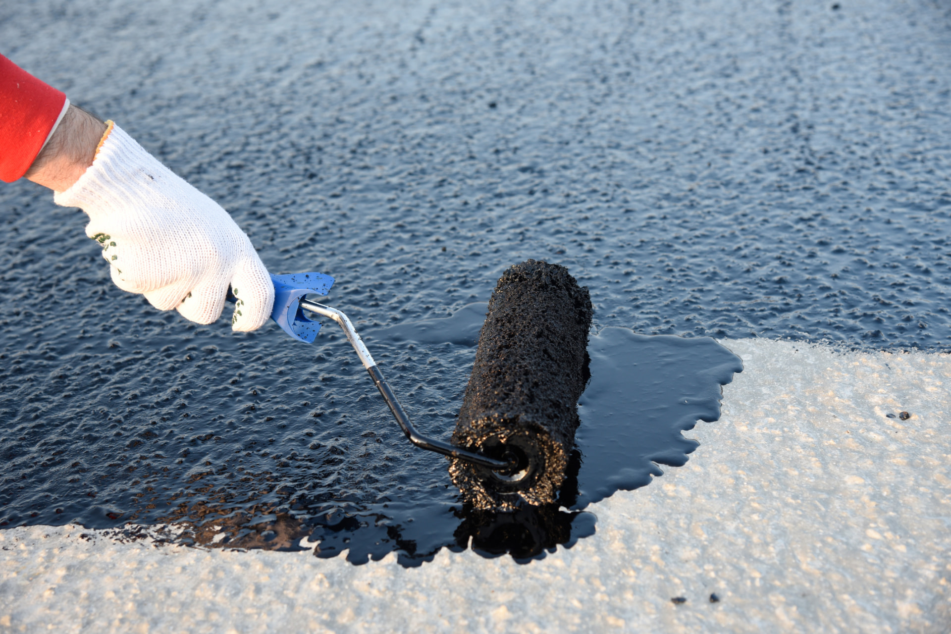Gloved worker rolling a pool of sealer across a faded asphalt surface