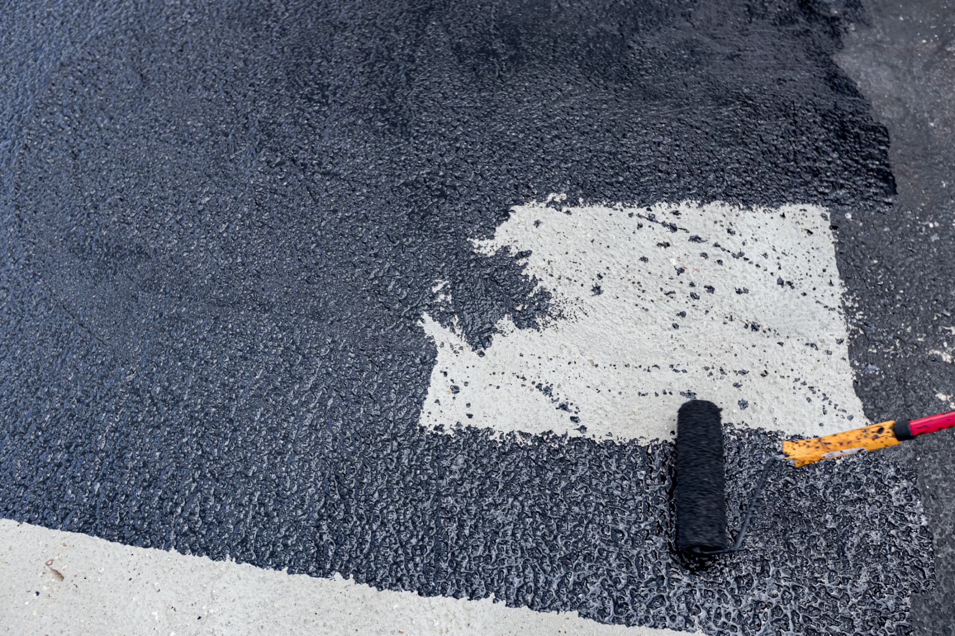Roller applying fresh sealcoat over asphalt beside a painted line