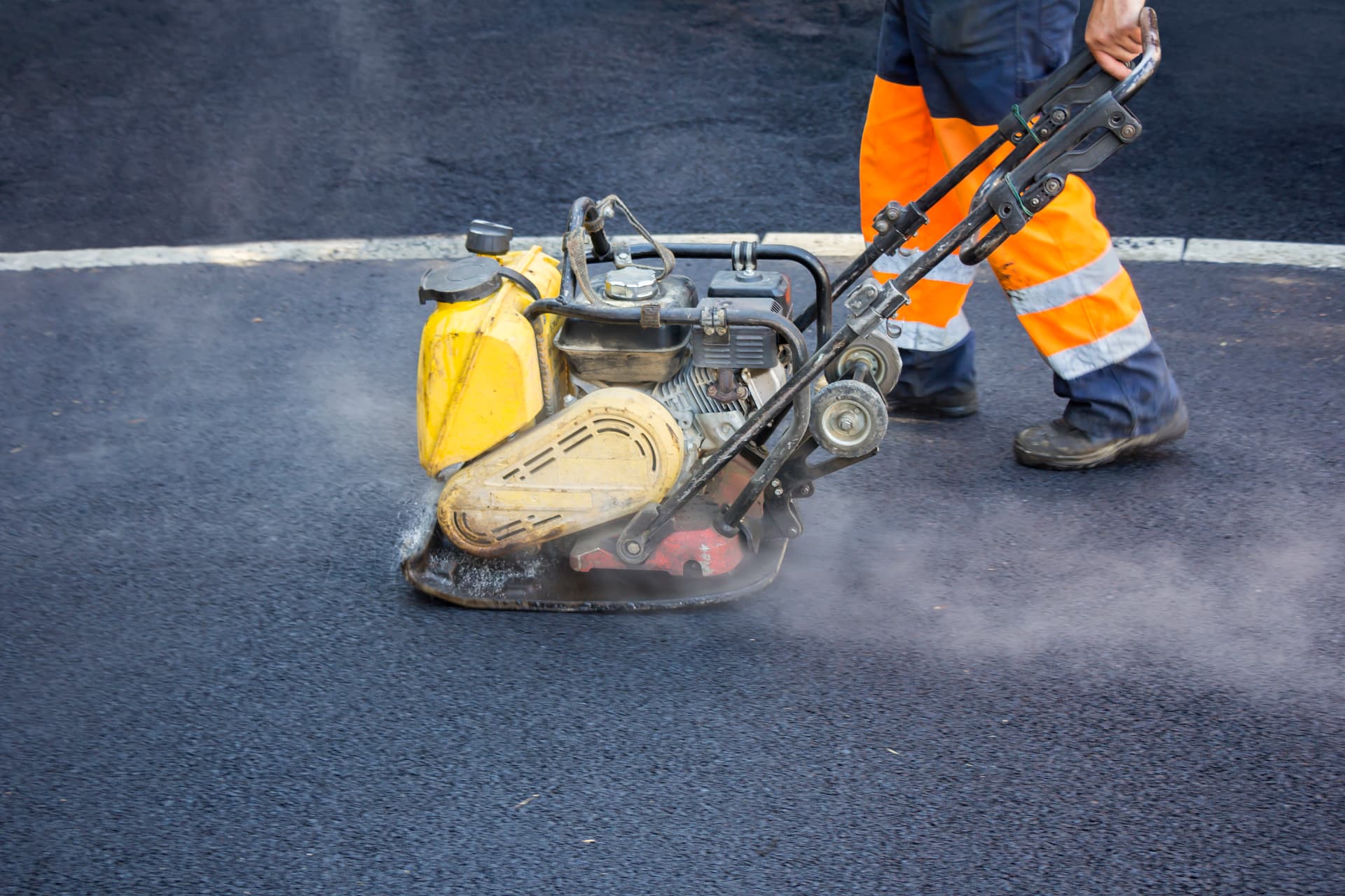 Worker compacting fresh hot-mix asphalt with a plate compactor on a paving project