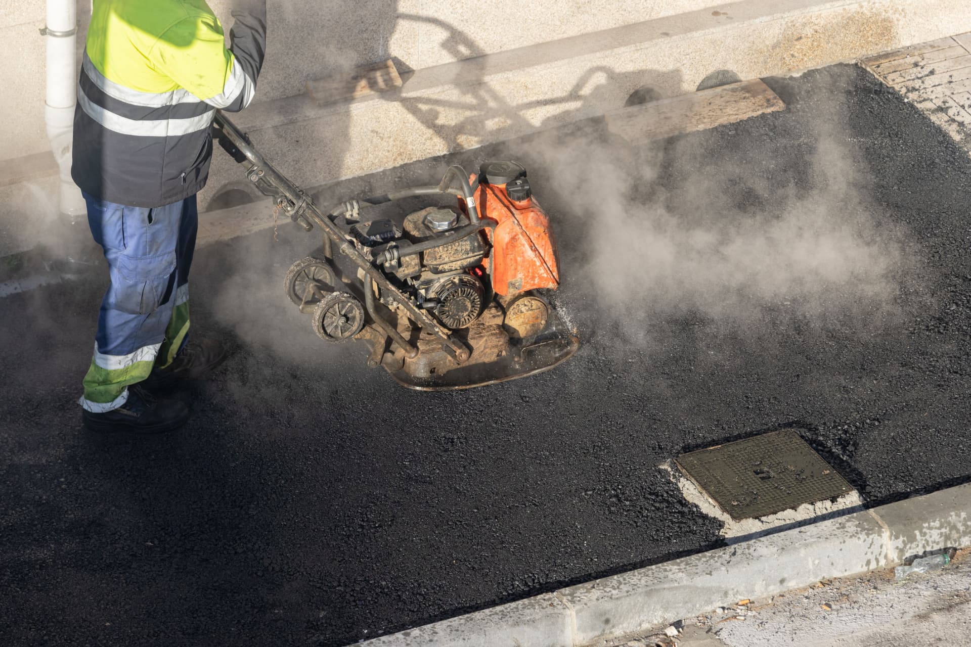 Worker compacting a steaming hot asphalt patch along a curb in Freehold, NJ