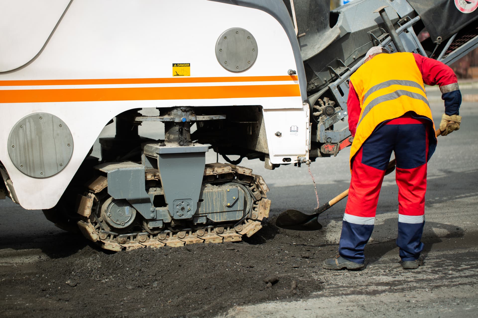 Worker shoveling asphalt beside a milling machine on a commercial paving project